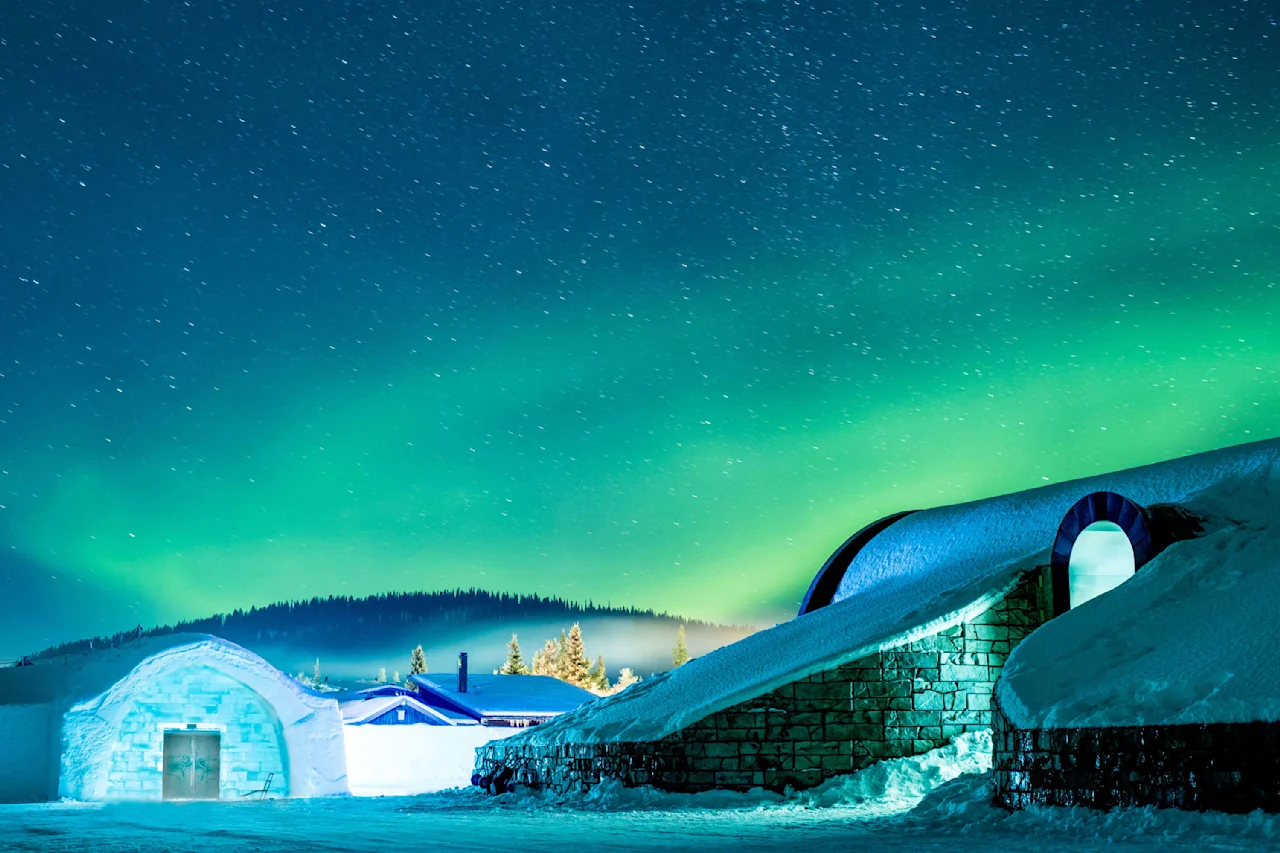 Snedækkede bygninger lyser op under et nordlys på en vinternat.