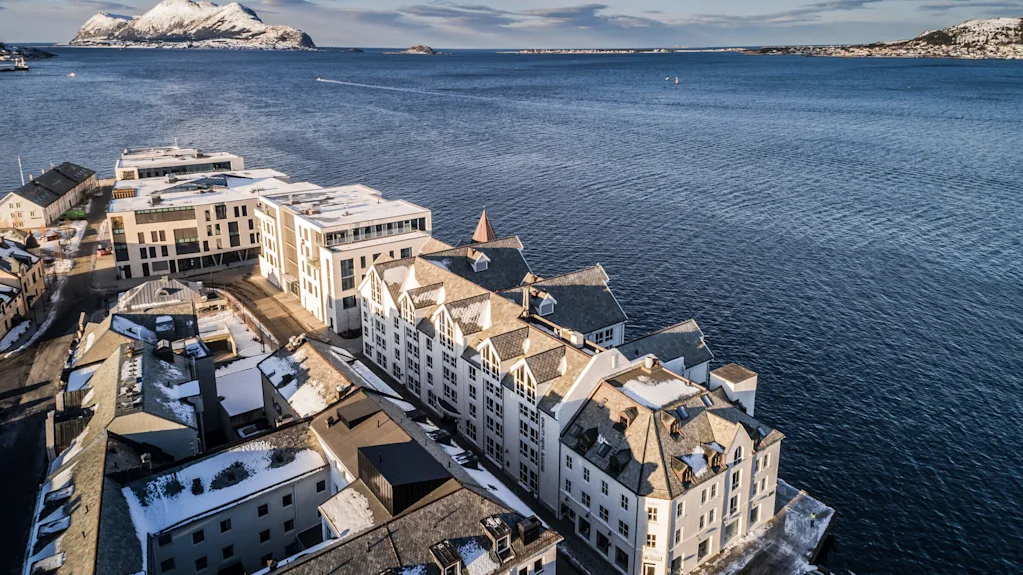 Snow-covered buildings line a coastal area facing a vast body of water. In the distance, snow-dusted mountains rise beneath a clear blue sky.