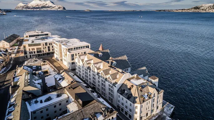 Snow-covered buildings line a coastal area facing a vast body of water. In the distance, snow-dusted mountains rise beneath a clear blue sky.