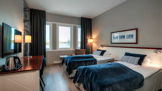 Twin beds with blue bedspreads are neatly arranged in a hotel room, featuring a wooden desk, flat-screen TV, and large windows with a sea view.