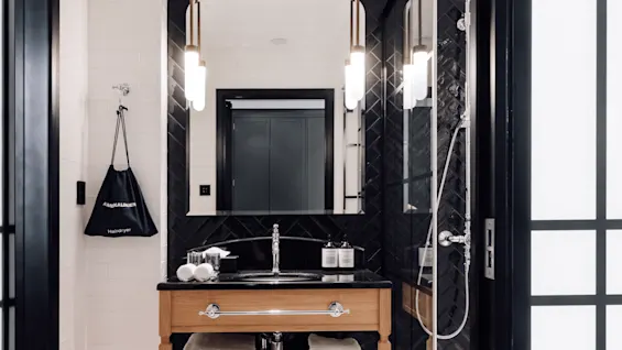 Vanity with a mirror and sink, underneath towels on a shelf, flanked by lamps, in a modern bathroom. A bag labeled “AMENALINEN Hairdryer” hangs on the wall.