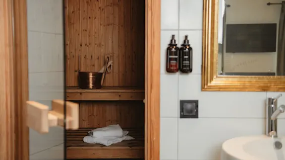 A warm and inviting sauna with wooden benches and a bucket, seen from a bathroom with a sink and a golden mirror.