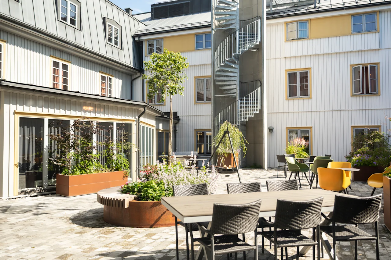 Utomhusgård med matbord och stolar på stenbelagd uteplats. Spiraltrappa leder upp längs byggnadens fasad. Krukväxter och små träd ger grönska till området.