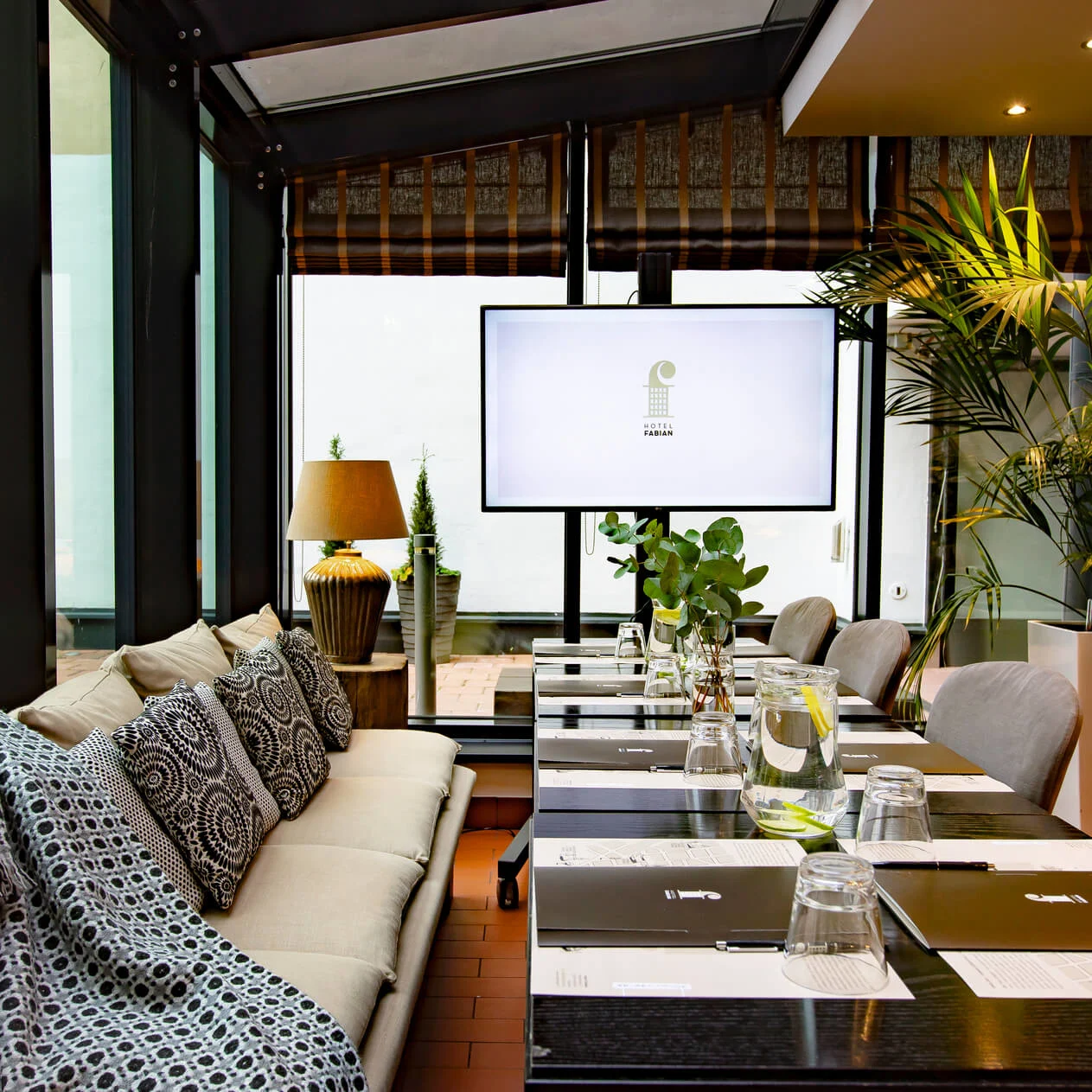 Et moderne møterom med et langbord dekket med glass og notatblokker. En stor skjerm viser "Hotel Fabian." Sofaer, dekorative puter og planter forbedrer omgivelsen.