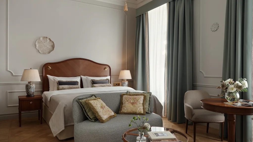 Ett prydligt arrangerat sovrum med en träbädd och kuddar samt lampor, omgiven av eleganta gardiner. En liten soffa och runt bord med blommor bidrar till rummets mysighet.