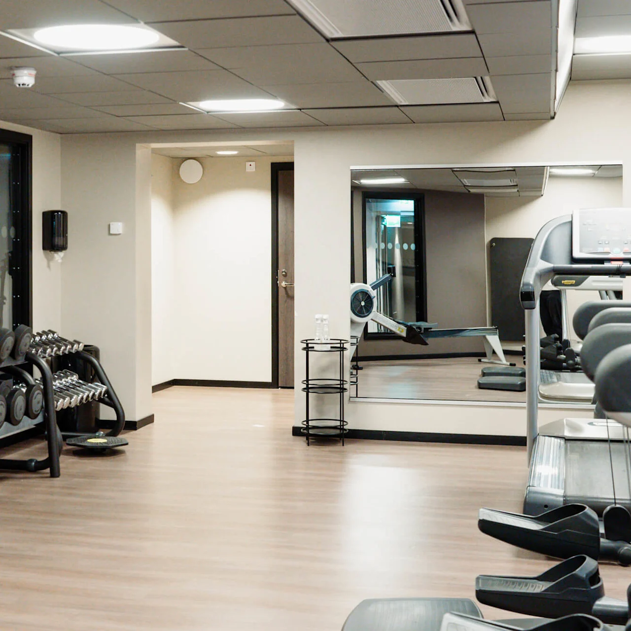 Et fitnesscenter med håndvægte på stativer, motionscykel og løbebånd i et veloplyst spejlrum med trægulv.