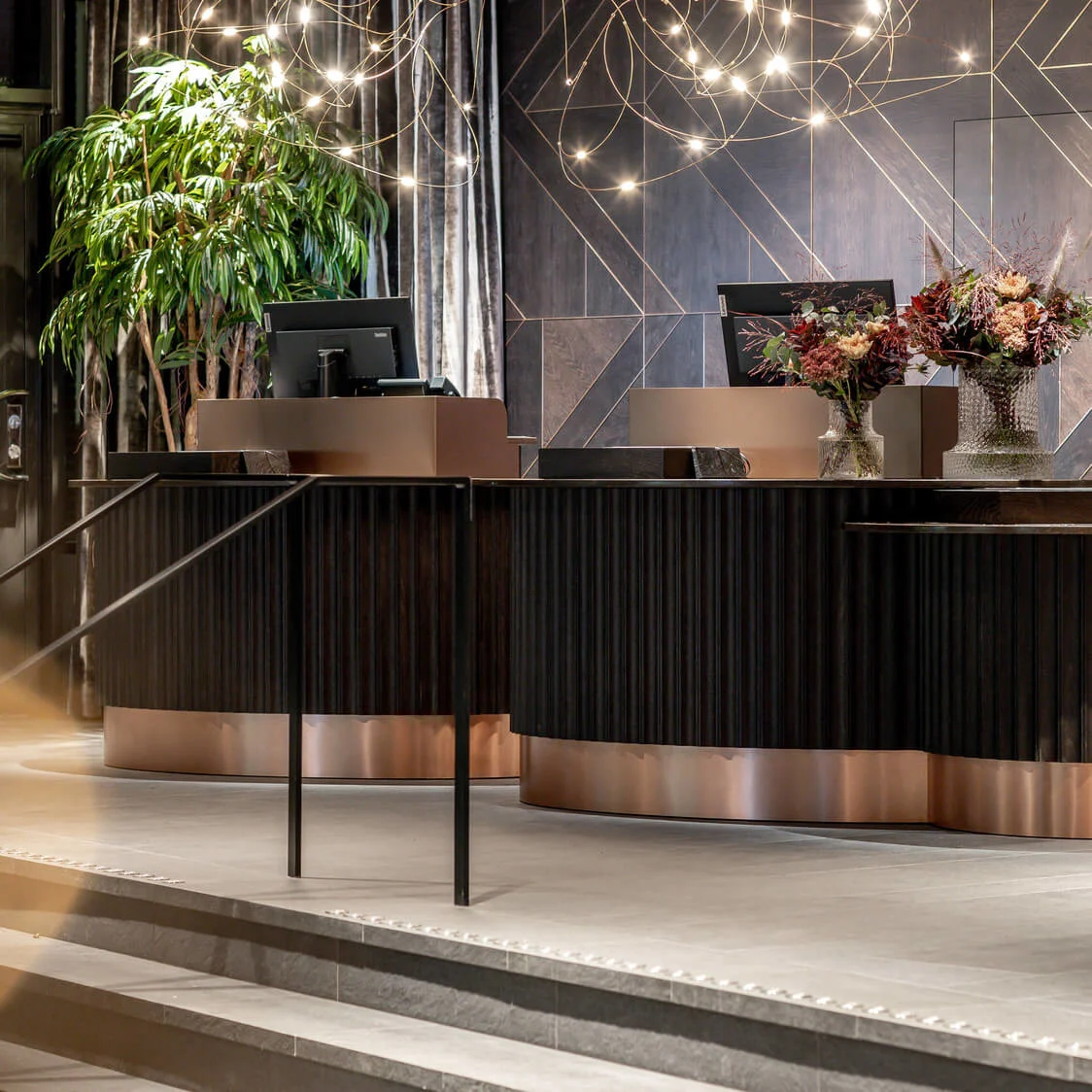 Receptiondisk med computere og blomsterarrangementer; oplyst af dekorative loftslamper, omgivet af geometrisk mønstret væg og potteplanter i et moderne, luksuriøst miljø.