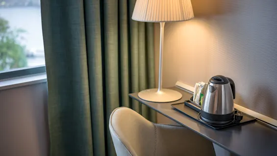 En kedel står på en bakke på et slankt sort bord ved siden af en beige stol. En tændt lampe oplyser miljøet ved siden af et vindue med grønne gardiner.
