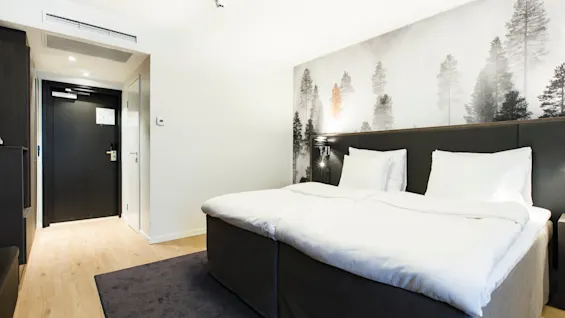 A neatly made bed with white linens stands in a modern hotel room, featuring a forest-themed mural on the wall, surrounded by light wooden flooring and a dark door.
