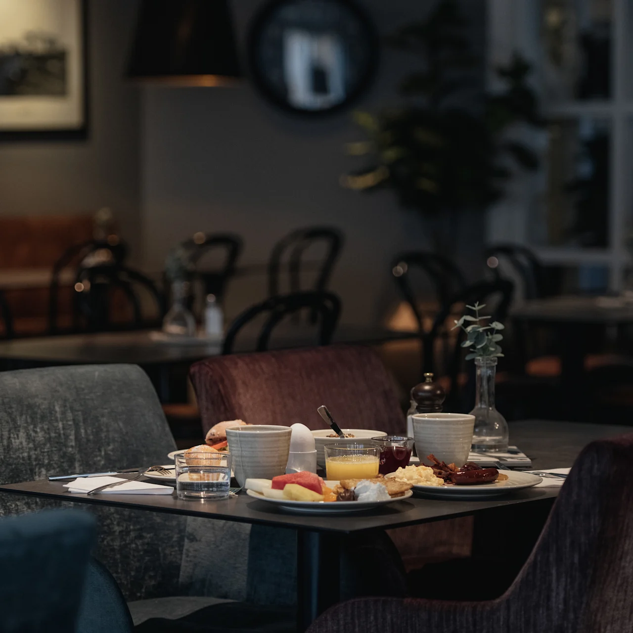 A dining table set with breakfast items, including juice, pastries, and coffee, sits in a cozy, dimly lit restaurant with neatly arranged chairs and framed photographs on the walls.