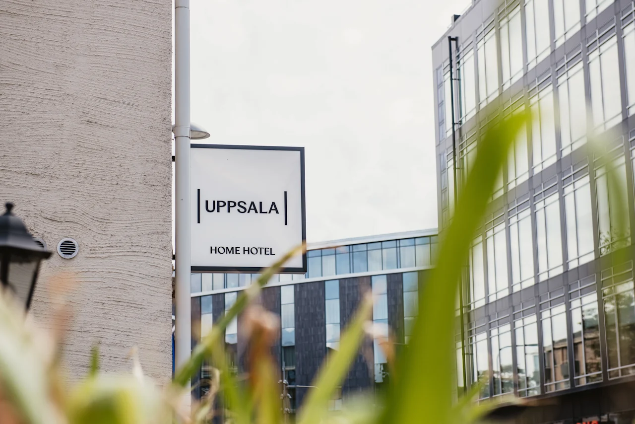 Exterior view of a modern building with a sign that reads UPPSALA HOME HOTEL, framed by green foliage.