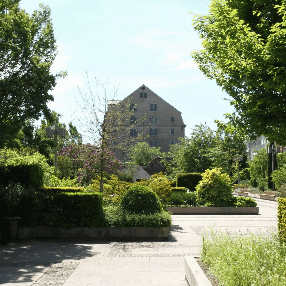 A large brick building stands amidst lush greenery, surrounded by trees, shrubs, and a pathway, under a clear blue sky.