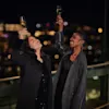 Two people hold champagne glasses aloft in celebration on a balcony, surrounded by city lights at night.