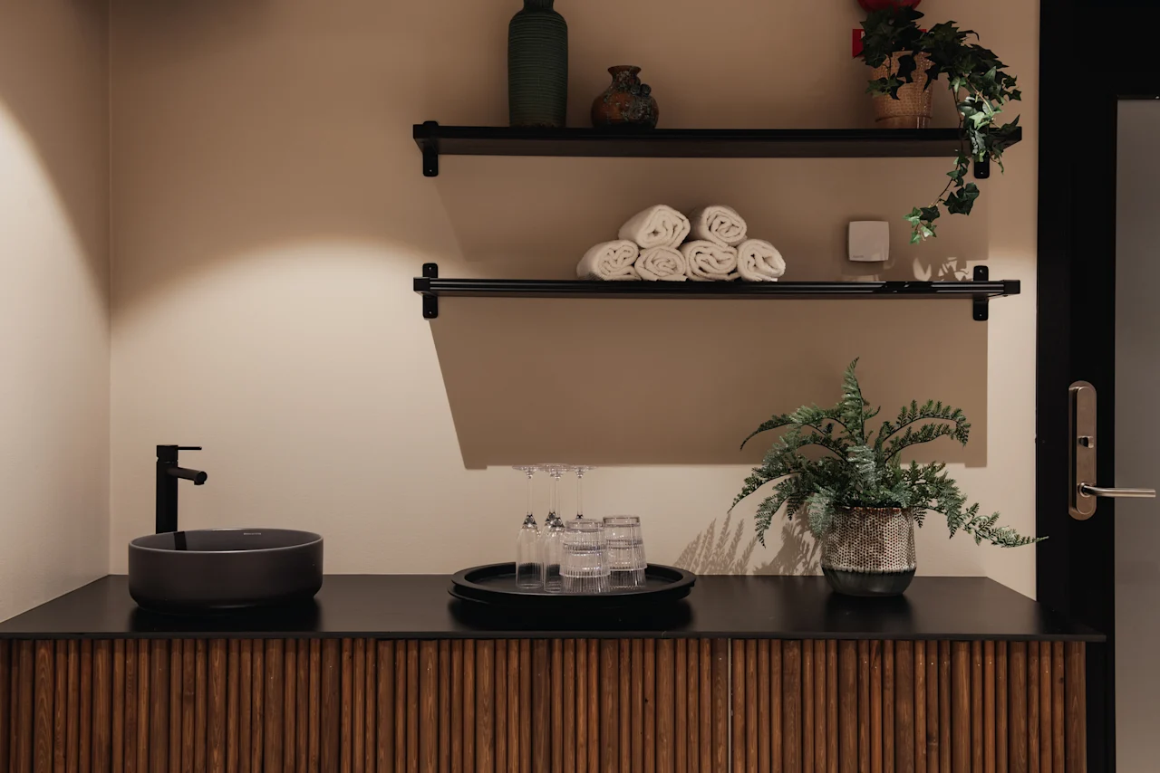 A stylish vanity area at Home Hotel Uppsala with a dark sink, rolled towels on shelves, and green plants, creating a serene atmosphere.