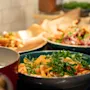 Danish: En blå skål indeholder en farverig salat med grøntsager og urter, omgivet af flere tallerkener med farverig mad på en køkkenbord, under varmt lys.