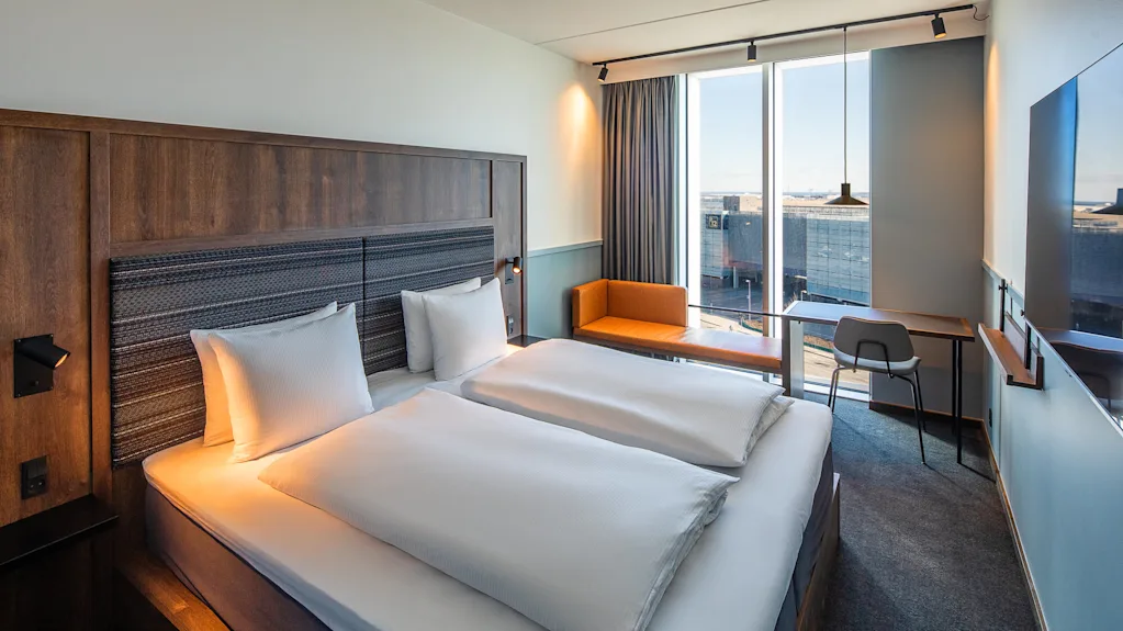 A hotel room contains two neatly made beds. Next to them, a wooden desk and chair sit by a large window offering a city view. Wall-mounted TV is present.