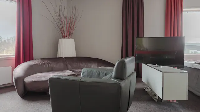 Moderne stue med en brun skinnsofa vendt mot en elegant TV på et hvitt stativ. Røde og mørke gardiner rammer inn store vinduer og tilfører varme til den nøytrale innredningen.