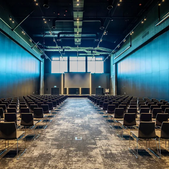 Rækker af tomme sorte stole vender mod en stor skærm i et rummeligt konferencelokale oplyst af loftlys. Høje vinduer giver naturligt lys.