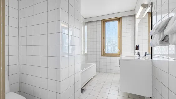 Lyst flislagt baderom med badekar, servant og et vindu som slipper inn naturlig dagslys.