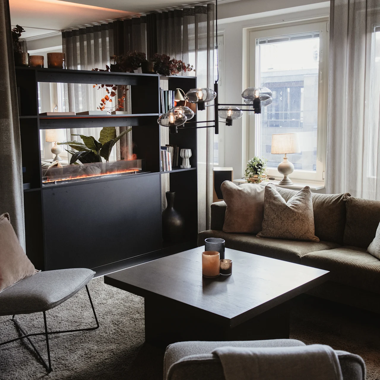 A cozy living room features a gray sofa adorned with pillows and a central black coffee table holding candles, bathed in soft light from large windows and modern light fixtures.