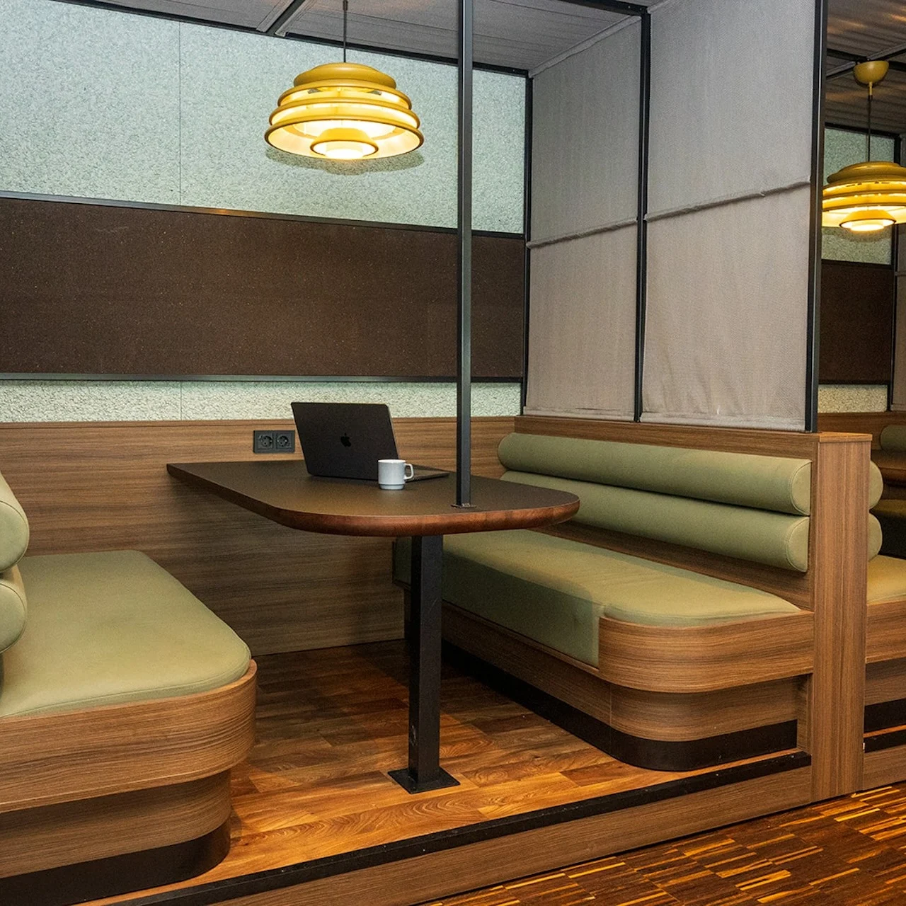 Cozy booth seating with a wooden table hosts a laptop and a mug in a warmly lit workspace. Modern pendant lighting and plush benches enhance the intimate office setting.