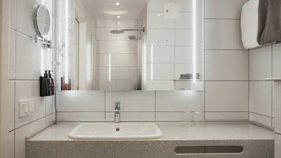 Moderni kylpyhuoneen pesuallasalue Home Hotel Victoriassa, jossa on suuri valaistu peili, joka heijastaa suihkua, valkoiset kaakeliseinät ja saippua-annostelijat.