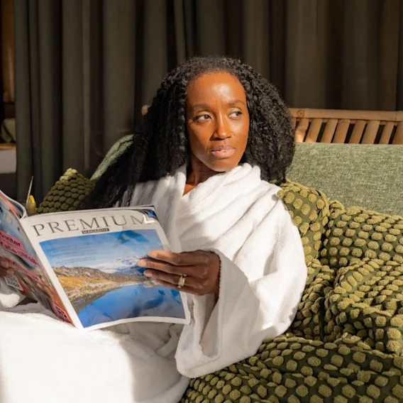 A person in a white robe reads a magazine titled "PREMIUM" while sitting on a green cushioned sofa, surrounded by soft, patterned pillows in a sunlit room.