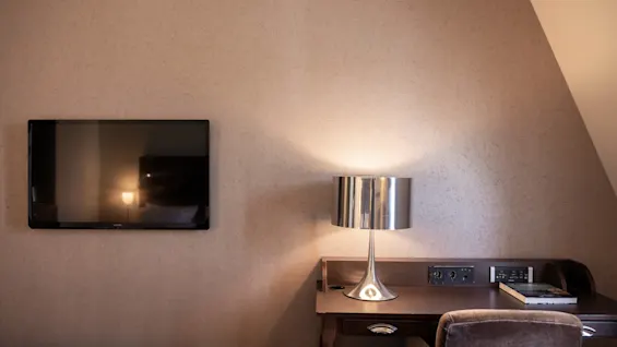 Et vægmonteret fjernsyn er slukket ved siden af et træskrivebord med en metallampe. En stol er skubbet ind under skrivebordet, der har en bog og stikkontakter i et hyggeligt rum.