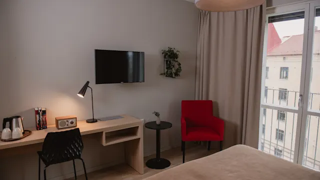 Ett prydligt hotellrum har ett skrivbord med lampa och stol, en TV på väggen, en röd stol bredvid ett litet bord och ett stort fönster med gardiner.