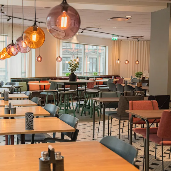 Tables and chairs are neatly arranged in a modern dining area, lit by spherical pendant lights. Large windows allow natural light, with patterned floor tiles adding elegance.