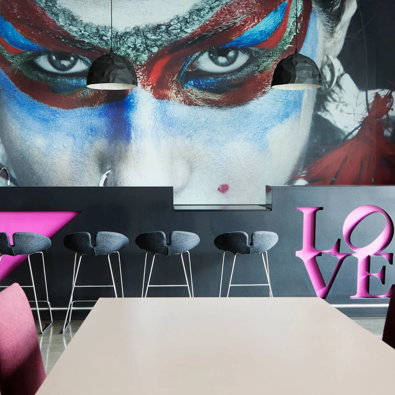 Et veggmaleri av et ansikt med fargerik maling omgir en bar med svarte stoler. Rosa bokstaver staver "LOVE". Hengende lys lyser opp scenen.