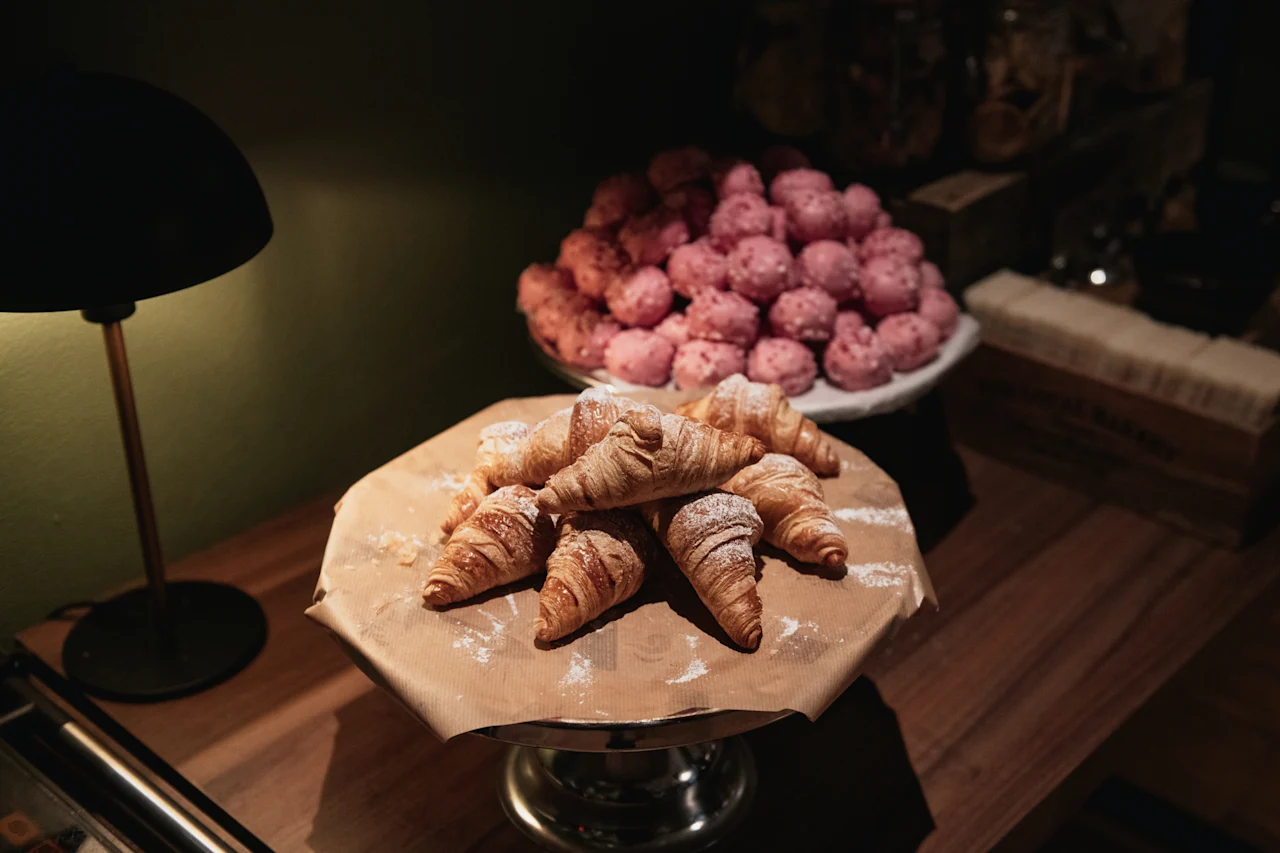 Vastaleivotut croissantit tomusokerilla pölytettynä telineellä, vaaleanpunaisten leivonnaisten kanssa taustalla.
