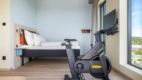 A modern hotel room featuring a comfortable bed and an exercise bike next to a large window, offering a view outside.