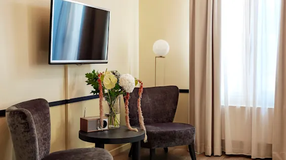 A wall-mounted TV is turned off above two dark chairs. A vase with flowers and a small radio sit on a round table. A window with curtains brightens the room.