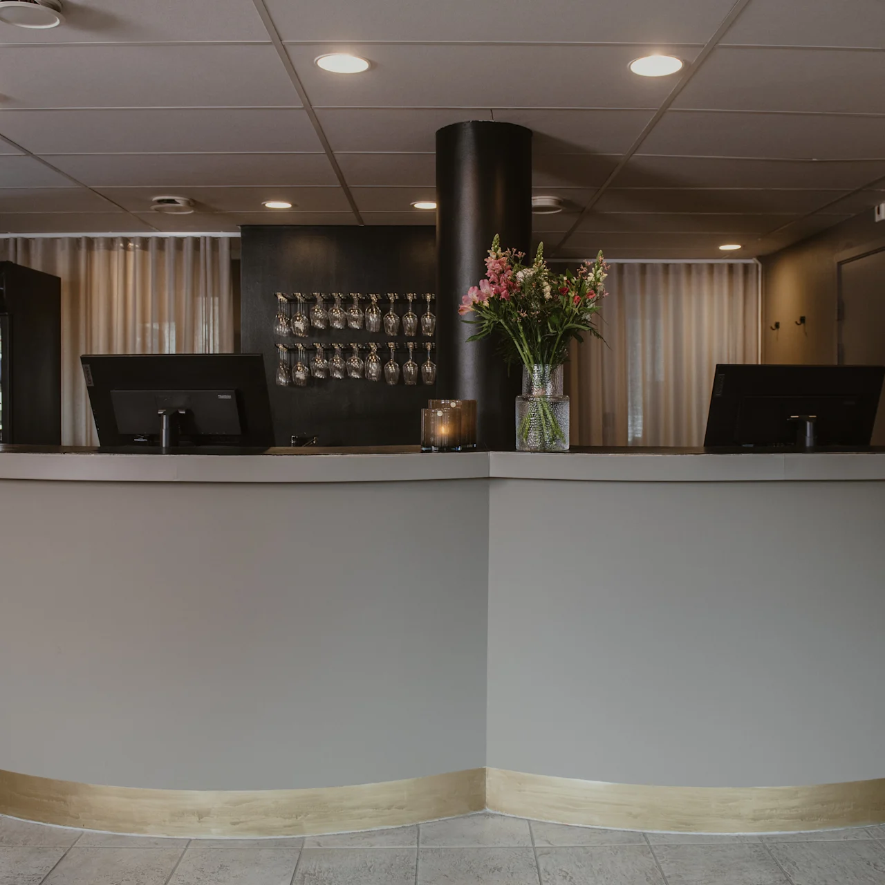 Reception desk displaying two monitors and a vase of flowers. A Carlsberg drink cooler stands nearby. Wine glasses are visible on a rack against the wall.