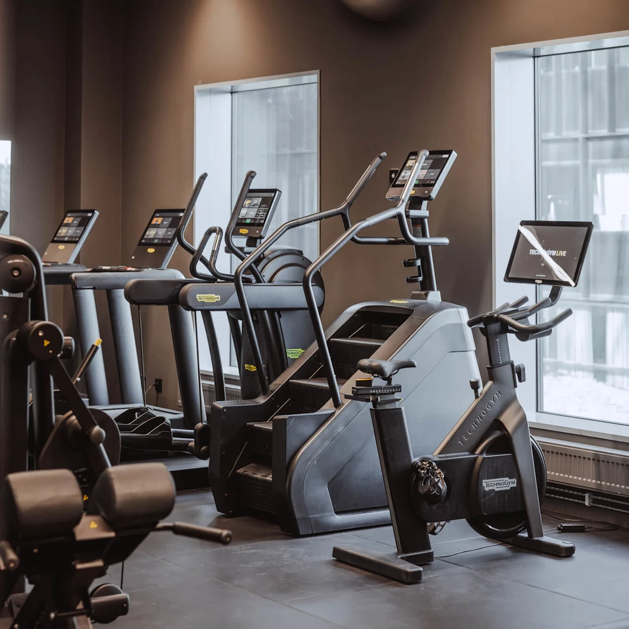Löpband och träningsmaskiner finns i ett modernt gym, vända mot fönster. Text på en skärm lyder: "TECHNOGYM LIVE". Annan utrustning är prydligt placerad på det mörka golvet.