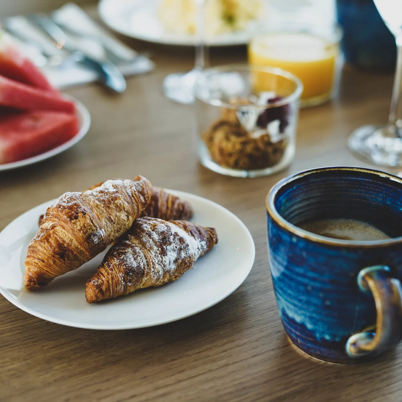 To croissanter ligger på en hvit tallerken ved siden av en blå kaffekopp på et trebord, omgitt av diverse frokostmat inkludert vannmelon, brød og appelsinjuice.
