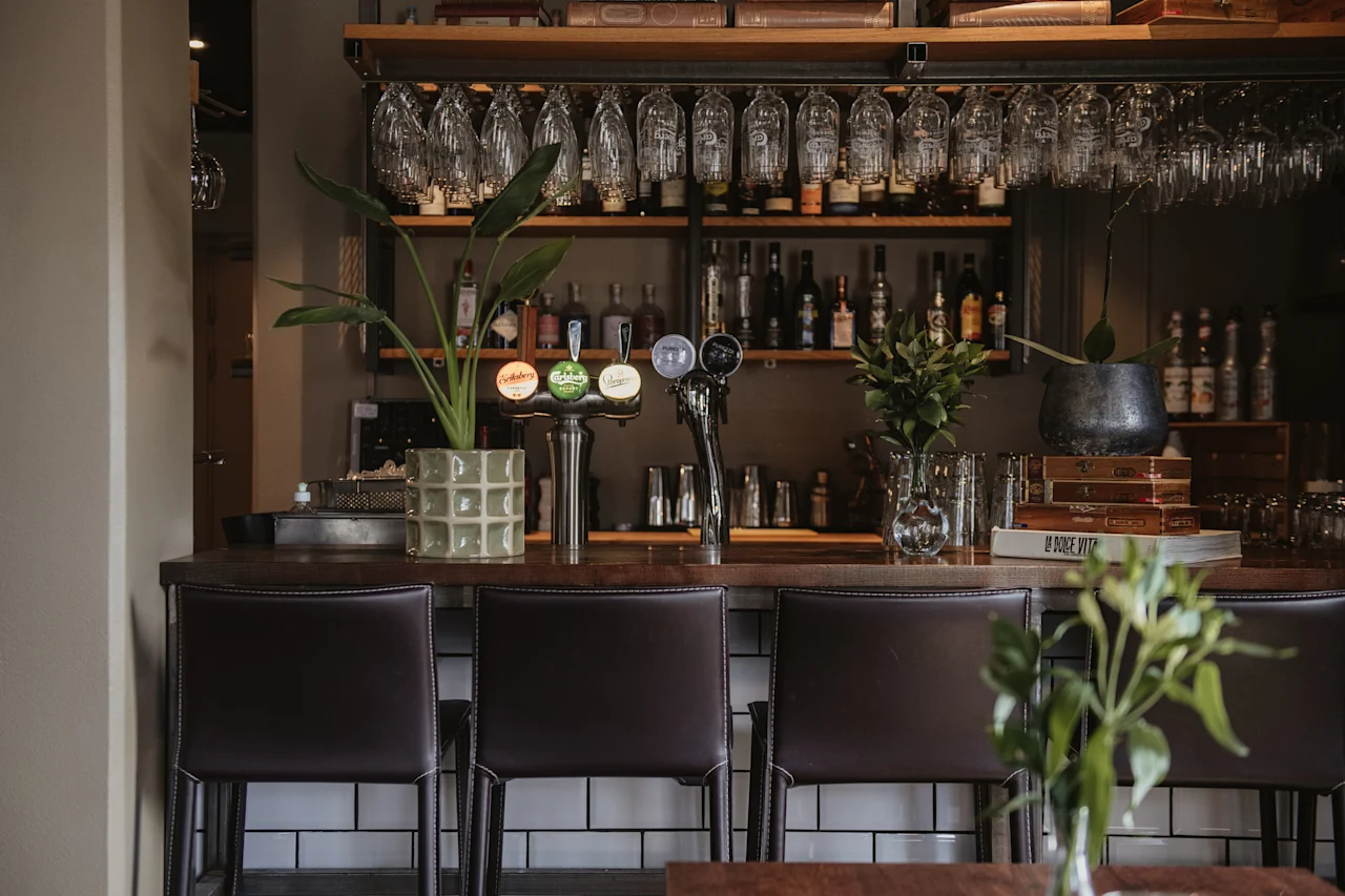En bar med mörka stolar, växter och hängande glas. Olika spritflaskor står på hyllorna. En liten vas med blad står på disken.