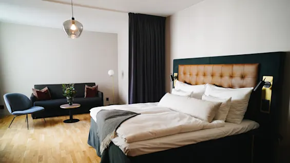 En sirligt redt seng med hvide lagner og en polstret sengegavl står i et rummeligt rum. I nærheden står en sofa med puder overfor et sofabord med en plante, under en hængende lampe.