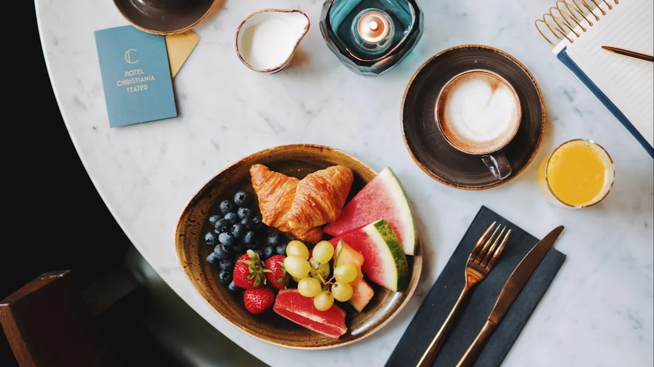 En frukostuppsättning på ett marmorbord med frukt, en croissant, kaffe, apelsinjuice och tända ljus. En anteckningsblock och HOTEL CHRISTIANIA TEATER-kort finns med.