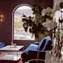 Mysig hotellounge med blå sammetssoffor, varm belysning, gröna växter och ett stort bågformat fönster.