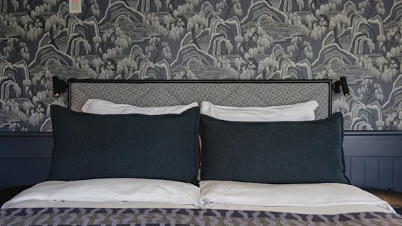 En mysig säng med mörkblå kuddar och en mönstrad filt, placerad mot en blå och vit landskapsmönstrad tapet.