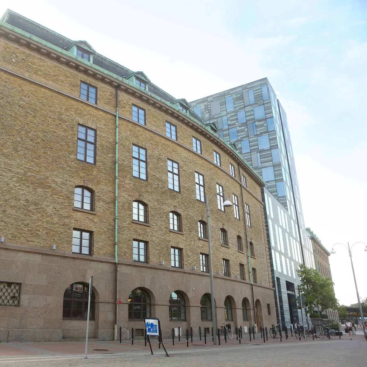 Suuri tiilirakennus, jossa on kaari-ikkunat, seisoo aurinkoisella kadunkulmalla. Kadun kyltti lukee "Åkerströmsgatan" jalankulkijoiden ylityskohdan vieressä.