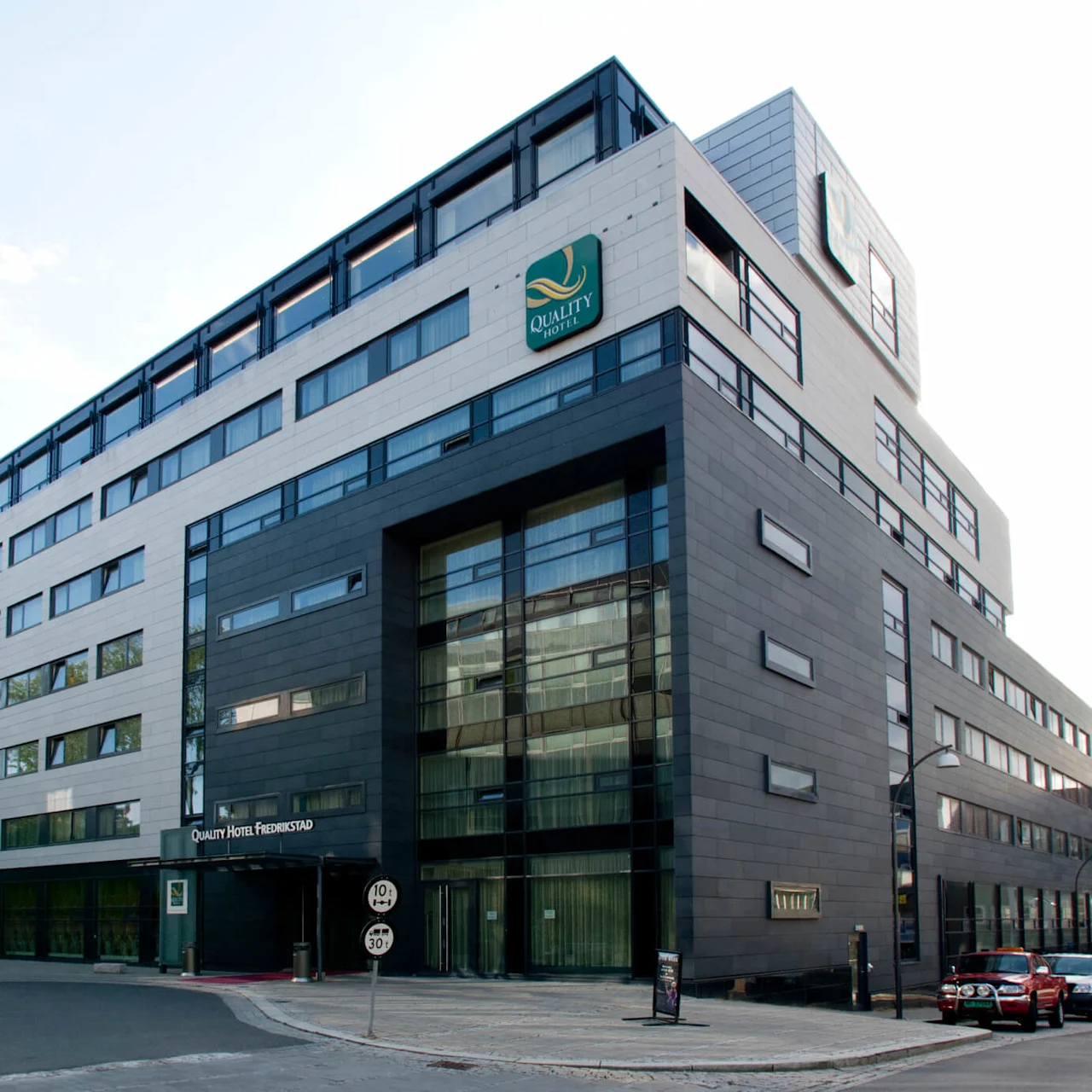 A modern hotel building stands with large windows and a mix of gray and white facade. Text: "Quality Hotel Fredrikstad." Situated on a quiet street with parked cars.