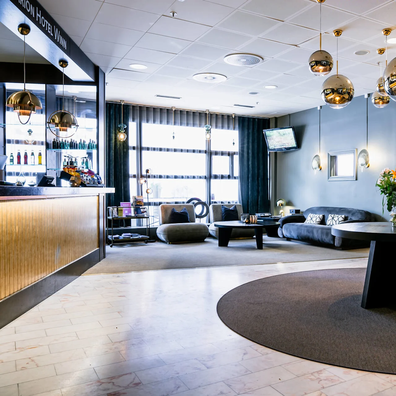 En hotellobby med moderne indretning har et rundt bord med blomster, hængende sfæriske lamper og et hyggeligt siddeområde. Tekst: "Clarion Hotel Winn" på receptionsdisken.