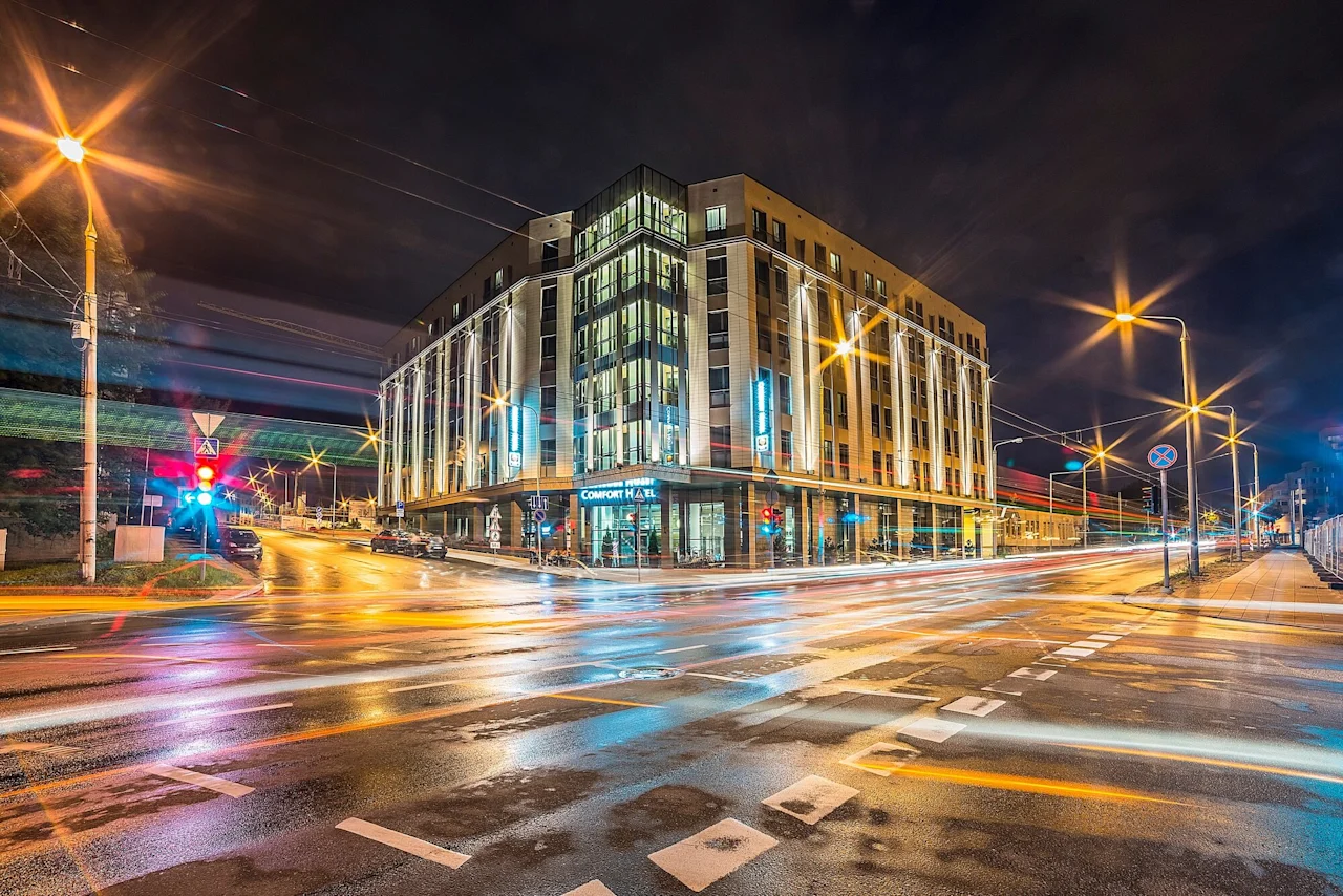 A modern hotel building with illuminated windows stands on a street corner at night, surrounded by light trails from passing vehicles. Text: "Comfort Hotel."