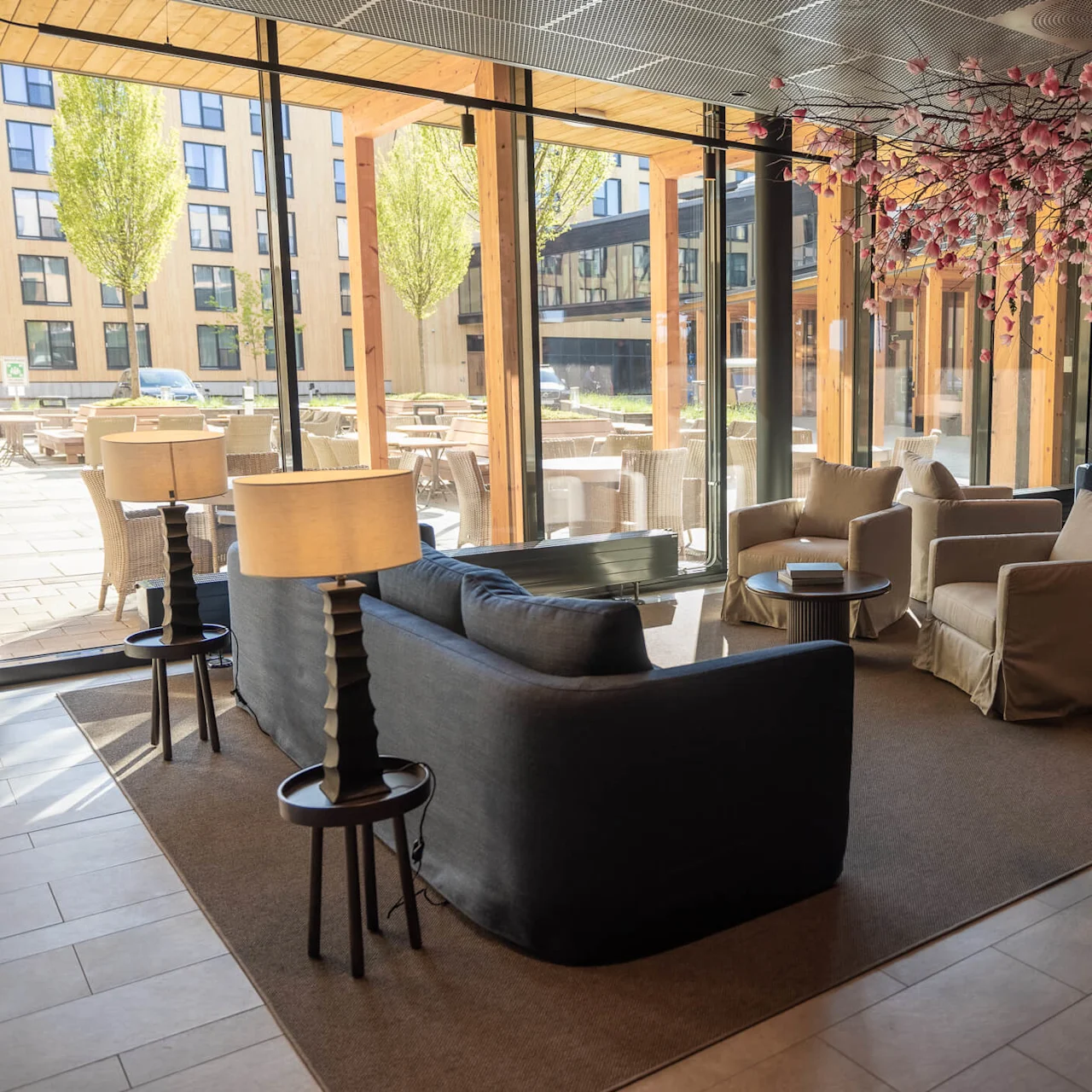 En modern lounge med soffor, stolar och ett dekorativt körsbärsträd ovanför. Stora fönster visar en uteservering och stadsmiljö.