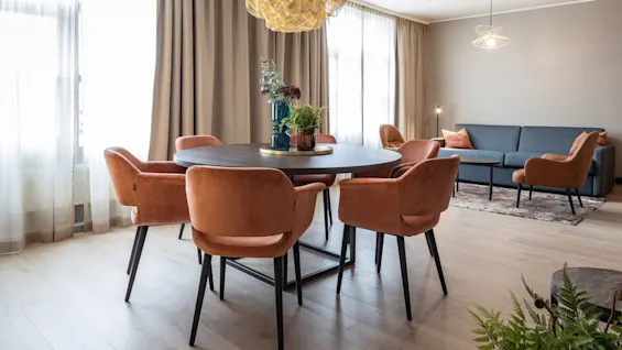 Spisebord omgivet af orange stole, pyntet med vaser, placeret i en moderne stue. Sofa og lænestole komplementerer rummet, oplyst af dekorative pendellamper.