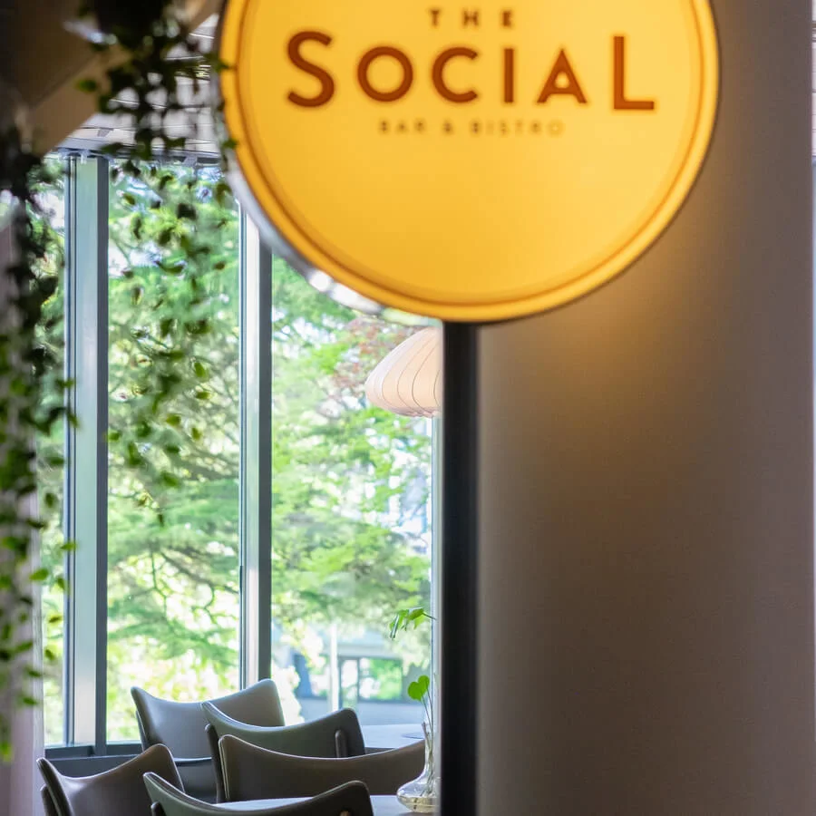 A round, yellow sign labeled "THE SOCIAL BAR & BISTRO" overlooks a cozy, green-filled dining area with modern chairs and large windows showing trees outside.