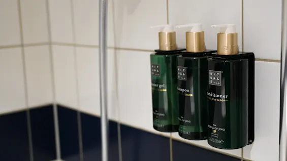 Tre gröna flaskor märkta "Rituals" är monterade på kaklad badrumsvägg. De är duschgel, schampo och balsam från "The Ritual of Jing"-kollektionen.